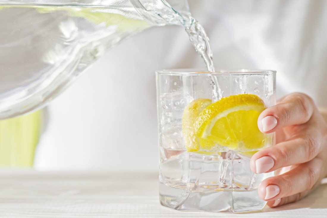 A person pours a pitcher of a water into a glass with a lemon slice in it